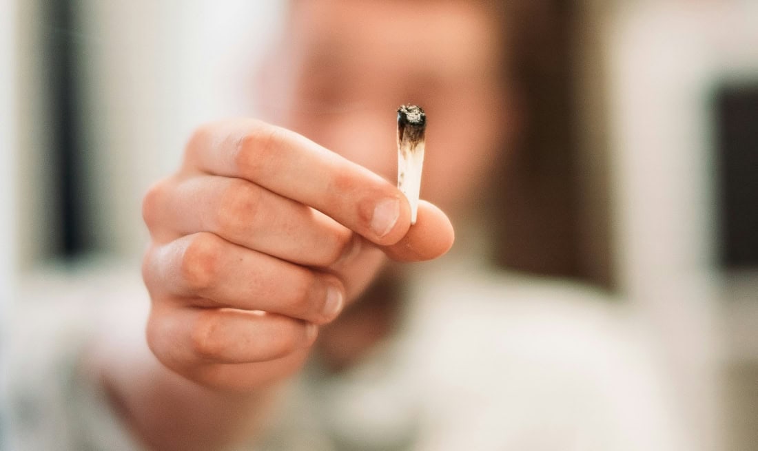 Person holds cigarette with ash in hand, focus on hand and cigarette.