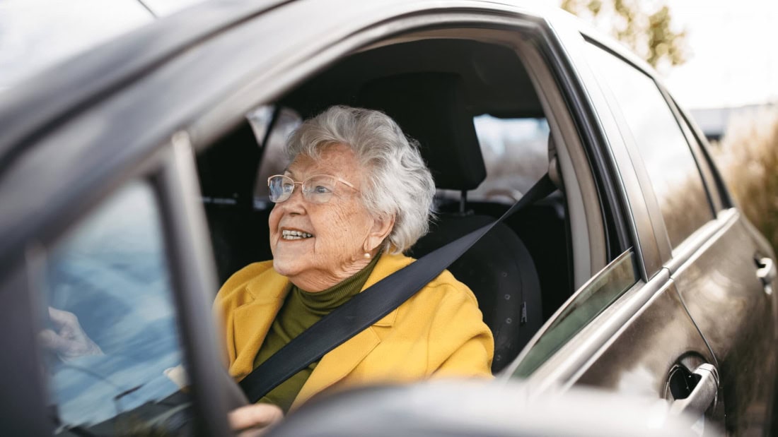 Oudere vrouw in auto tijdens rijbewijskeuring, glimlachend en ontspannen.
